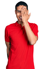 Fototapeta premium Young hispanic man wearing casual red t shirt covering one eye with hand, confident smile on face and surprise emotion.