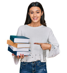 Young hispanic girl holding a pile of books smiling happy pointing with hand and finger