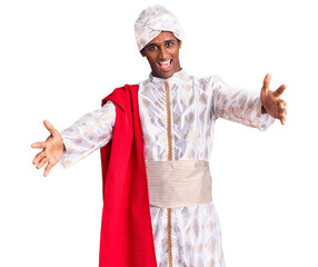 African handsome man wearing tradition sherwani saree clothes looking at the camera smiling with open arms for hug. cheerful expression embracing happiness.