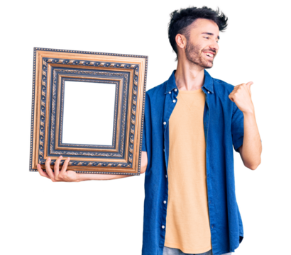 Young hispanic man holding empty frame pointing thumb up to the side smiling happy with open mouth
