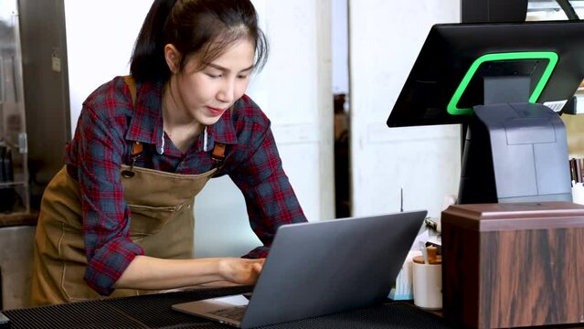 Closeup Asian cafe small business owner female standing in counter making order selling transaction checking turnover profit management using laptop technology, barista waitress worker happy smiling