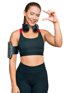 Beautiful blonde woman wearing gym clothes and using headphones smiling and confident gesturing with hand doing small size sign with fingers looking and the camera. measure concept.