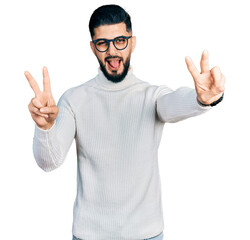 Young arab man with beard wearing elegant turtleneck sweater and glasses smiling with tongue out showing fingers of both hands doing victory sign. number two.