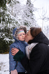 Mom with daughter in winter snowy forest. Mom kisses her daughter and warms her hands with her breath. Young girl with her daughter walking in the forest in winter.