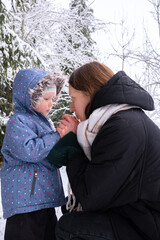 Mom with daughter in winter snowy forest. Mom kisses her daughter and warms her hands with her breath. Young girl with her daughter walking in the forest in winter.