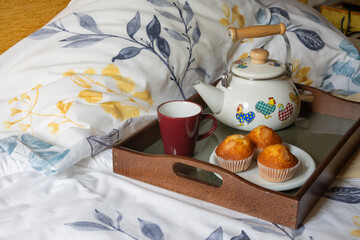 Top view of tray with colorful teapot, cup and muffins, on a bed with sheets and pillows with drawings of branches, yellow background, horizontal, with copy space