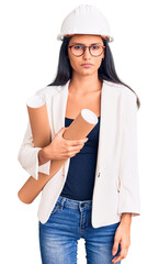 Young beautiful latin girl wearing architect hardhat holding blueprints thinking attitude and sober expression looking self confident