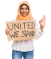 Young brunette arab woman holding united we stand banner doing ok sign with fingers, smiling...