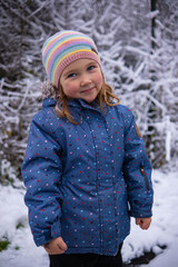 Little beautiful girl in winter clothes standing alone in the middle of a snowy forest. Girl in jacket with fur hood posing in winter forest. Shrubs and trees are covered with snow