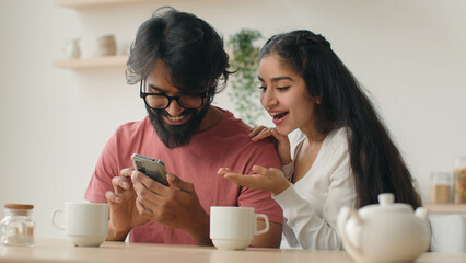 Excited multiethnic family Arabian indian man and woman looking at mobile phone screen at home kitchen happy couple girl and guy make order winning achievement shopping online with smartphone laughing