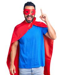 Young hispanic man wearing super hero costume pointing finger up with successful idea. exited and happy. number one.