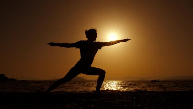 Woman enjoy warrior pose at sunset. A view of woman silhouette in warrior pose stand on the evening shore during summer vacation.