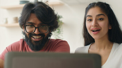 Happy Indian Arabian couple use laptop in kitchen smiling diverse man husband and woman wife surfing website watch funny family photos shopping at home laughing have fun talk with friends online laugh