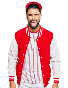 Young man with beard wearing baseball uniform sticking tongue out happy with funny expression. emotion concept.