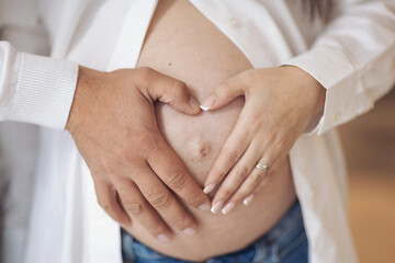 A couple forms loving heart on pregnant belly.Pregnant woman in white dress and her husband...