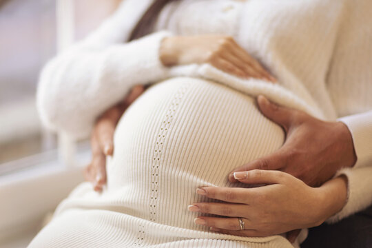 Pregnant woman in white dress and her husband indoors, closeup. Man touching his pregnant wife's belly at home. pregnancy expectation concept.