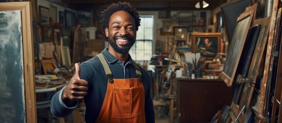 An African American painter in his studio, holding an empty frame, smiles happily and gives a thumbs up, expressing excellence and approval.