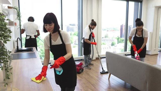 Cleaning service team at work in kitchen in private home. Caucasian janitor in apron cleaning table with detergent and gloves while her multiethnic colleague wiping floor, windows and vacuuming sofa.