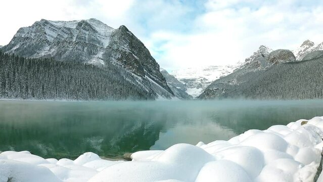 Winter at misty lake natural background footage. Cold icy moment. fluffy white snow front green emerald lake mountain scenery reflexed in clear water surface with cloudy blue sky at background.  