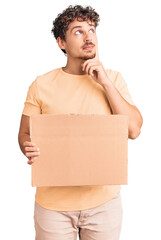 Young handsome man with curly hair holding blank cardboard empty banner serious face thinking about...