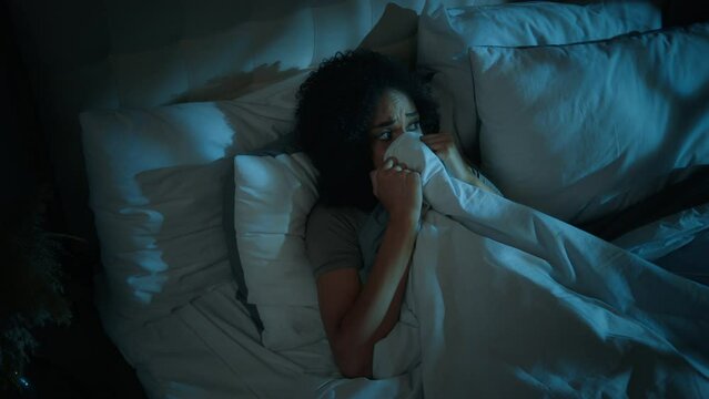 Scared frightened African American woman hiding under blanket bed nightmare sleep disorder bad dream disturbed afraid teenager girl in bedroom late night dark home nighttime fear scary monster phobia