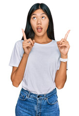 Beautiful young asian woman wearing casual white t shirt amazed and surprised looking up and pointing with fingers and raised arms.