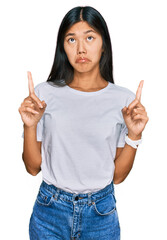 Beautiful young asian woman wearing casual white t shirt pointing up looking sad and upset, indicating direction with fingers, unhappy and depressed.