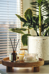 Apartment decoration with aroma reed diffuser, burning aromatic candles on wooden tray. Hygge, home aromatherapy, relaxation concept. Green plants on background. Bali oriental style in modern interior