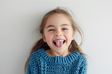 A cute Caucasian girl in a blue knitted sweater against a white wall with a happy and funny face, smiling and sticking out her tongue