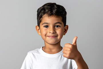A small Hispanic boy in a white T-shirt makes a happy hand gesture with his thumbs up. The approving expression on his face as he looks at the camera indicates success