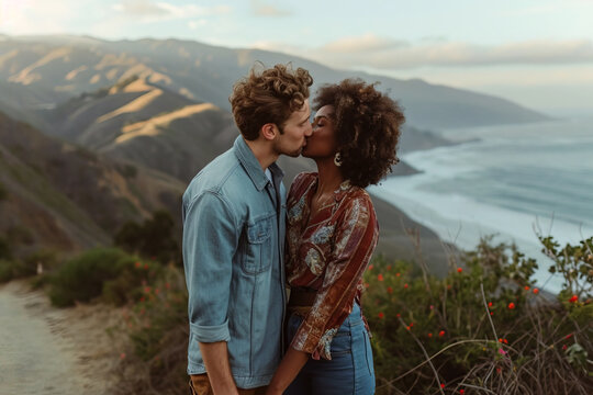 An authentic interracial couple, dressed casually, kiss tenderly, facing each other and holding hands, overlooking the ocean and mountains behind them