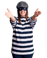 Young beautiful brunette woman wearing burglar mask looking at the camera smiling with open arms...