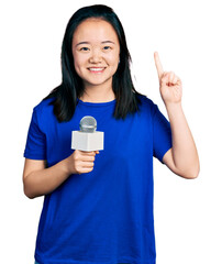 Young chinese woman holding reporter microphone smiling with an idea or question pointing finger with happy face, number one