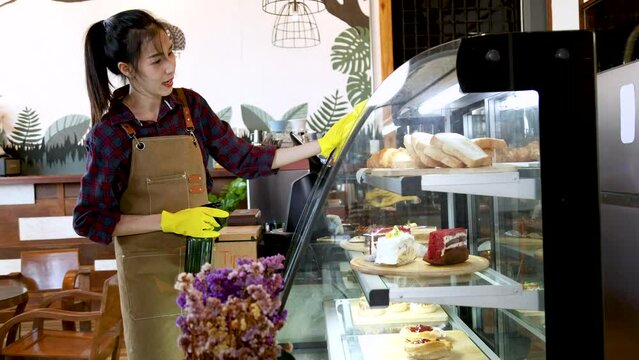 Asian barista waitress worker female wearing apron in cafe restaurant wearing gloves wipe and clean bakery cabinet preparing to open cafe shop, small business entrepreneur start-up multitask lifestyle