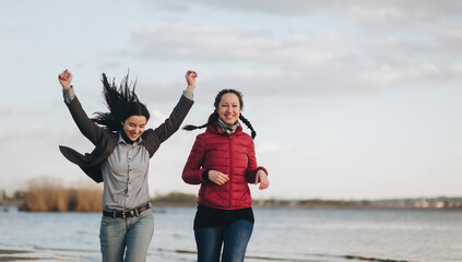 Two girlfriends in blue jeans, a brown and red jackets and a gray shirt run along the river bank in the evening of sunset and laugh with joy. The concept of a spring walk outdoors. - obrazy, fototapety, plakaty