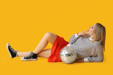 Beautiful thoughtful young woman with disco ball lying on yellow background