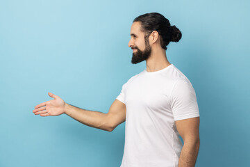 Side view of man with beard wearing white T-shirt standing and looking at camera with toothy smile and giving hand to greeting or handshake. Indoor studio shot isolated on blue background.