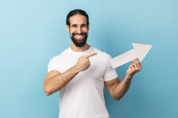 Portrait of smiling positive man with beard wearing white T-shirt holding white arrow and pointing...