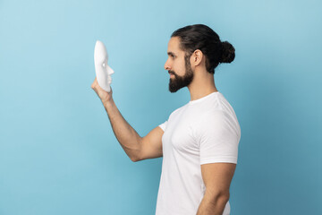 Side view of man with beard wearing white T-shirt holding and looking at white mask with attentive...