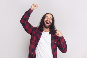Cheerful triumphing man with long curly hair in checkered red shirt achieves victory, raises clenched fists with triumph, rejoices winning prize. Indoor studio shot isolated on gray background. - obrazy, fototapety, plakaty