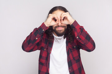 Portrait of bearded man with long curly hair in checkered red shirt making glasses shape with...