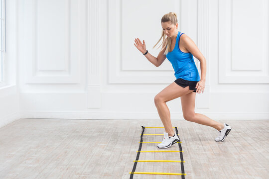 Workout and sport concept. Full length of sporty powerful blonde woman with ponytail in black shorts and blue top training on agility ladder drills in gym. Indoor studio shot.