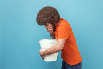 Side view of sick unhealthy man with Afro hairstyle wearing orange T-shirt standing with white bucket, vomiting, suffering bad feelings, discomfort. Indoor studio shot isolated on blue background. - obrazy, fototapety, plakaty