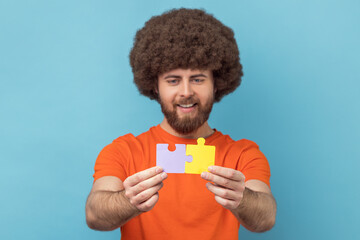 Portrait of man with Afro hairstyle holding two puzzle parts and smiling joyfully, ready to connect...