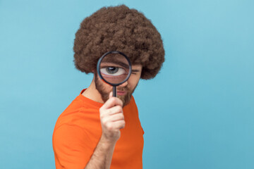 Funny man with Afro hairstyle wearing orange T-shirt standing, holding magnifying glass and looking...