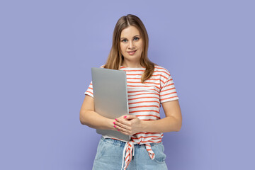 Portrait of cheerful happy optimistic blond woman wearing striped T-shirt standing with closed laptop, finishing her work, being satisfied. Indoor studio shot isolated on purple background.