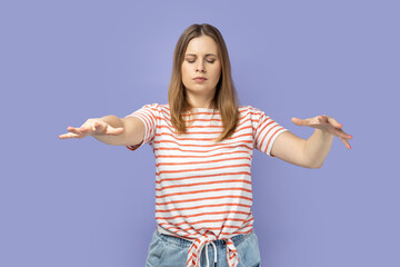 Vision problems. Blind disoriented lonely blond woman wearing striped T-shirt walking with closed eyes, raising hands to search lost road in darkness. Indoor studio shot isolated on purple background.