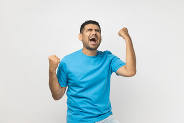 Portrait of unshaven man wearing blue T- shirt standing clenches fists with positive expression, rejoices wonderful news, smiles happily. Indoor studio shot isolated on gray background.