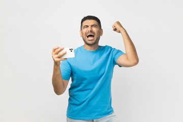 Portrait of extremely happy unshaven man gamer wearing blue T- shirt standing with mobile phone in...