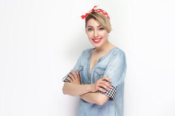 Portrait of happy joyful beautiful blonde woman wearing blue denim shirt and red headband standing with crossed arms, looking smiling at camera. Indoor studio shot isolated on gray background.